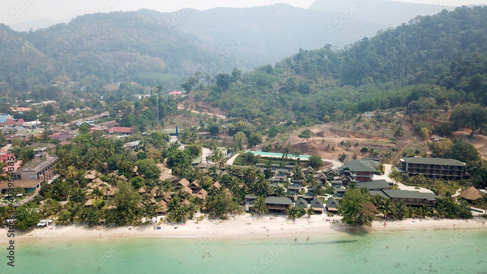 Green Chang island is covered in smoke from a fire in Cambodia. Green trees, palm trees and hills are covered with clouds of smog. Hotels stand on a cliff in front of the sea. Turquoise water.
