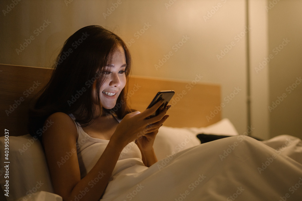 Smiling beautiful lady in white t-shirt on the bed using smartphone in the room Smiling beautiful lady in white t-shirt on the bed using smartphone in the room