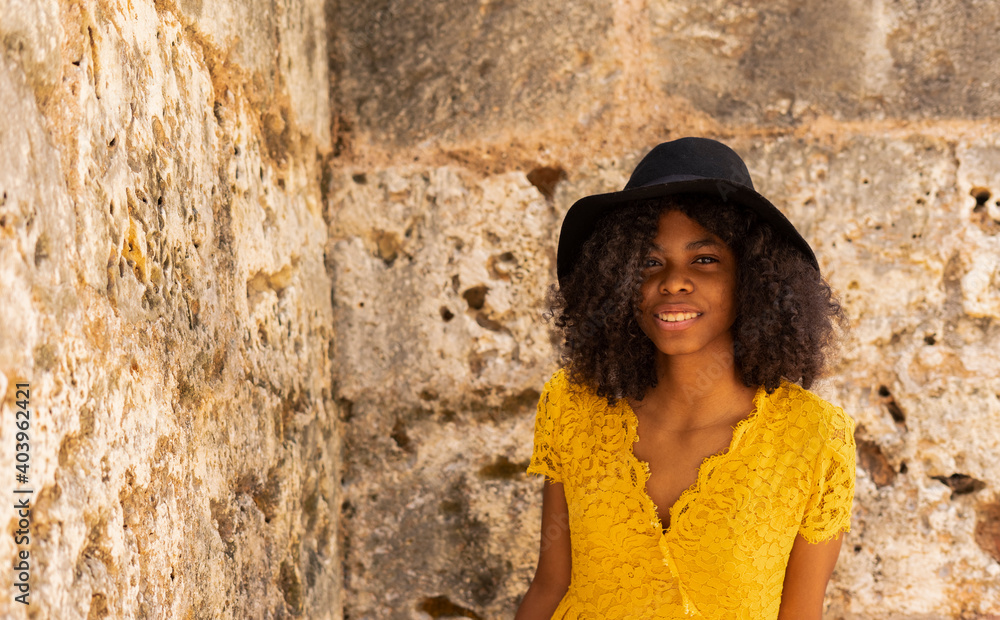 Young black woman with curly hair, wearing yellow dress and black hat, with styles and attitude, in front of a stone wall