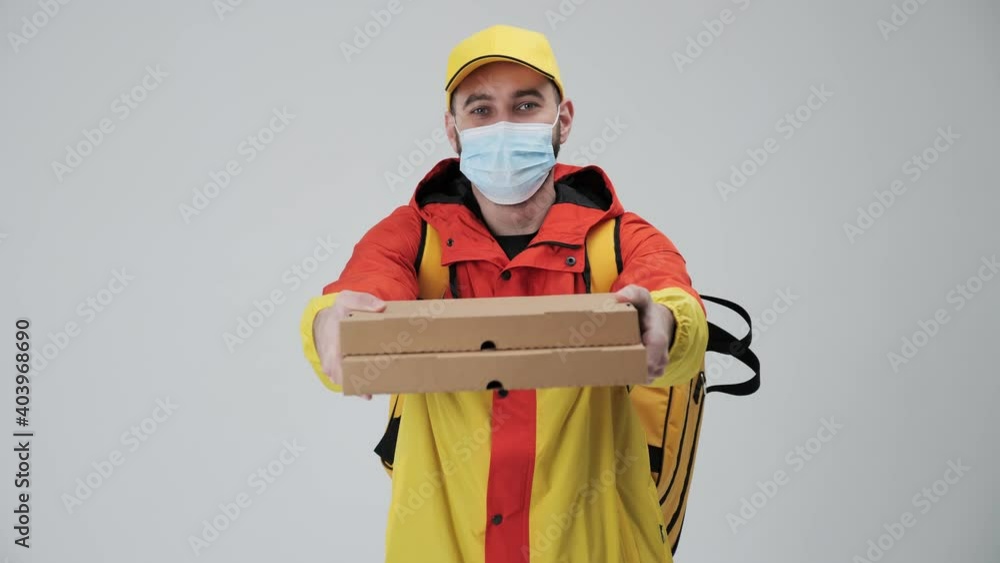 Bearded smiley delivery man holding pizza giving the order wearing red yellow uniform and cap wearing mask during covid-19 pandemic in studio on white background. Bearded smiley delivery man holding pizza giving the order wearing red yellow uniform and cap wearing mask during covid-19 pandemic in studio on white background.