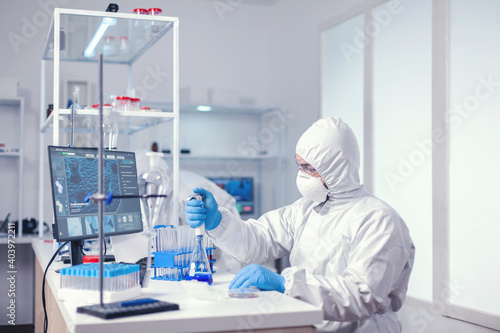 Experienced biologist dressed in ppe holding pipette with blue solution. Chemist in modern laboratory doing research using dispenser during global epidemic with covid-19.