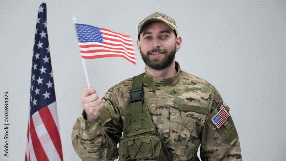 Cheerful soldier in a military uniform waves the American flag smiling looks to the camera near the American flag in studio on white background. Cheerful soldier in a military uniform waves the American flag smiling looks to the camera near the American flag in studio on white background.