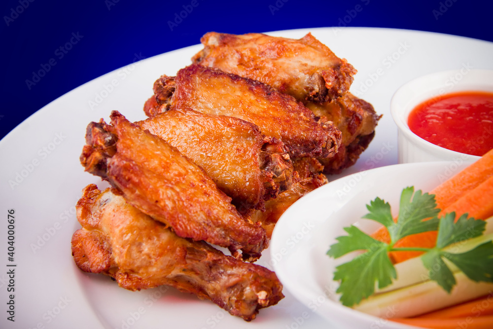 Roasted chicken wings with sticks carrots and celery and dipping sauce on white plate on blue background