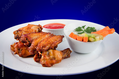 Roasted chicken wings with sticks carrots and celery and dipping sauce on white plate on blue background