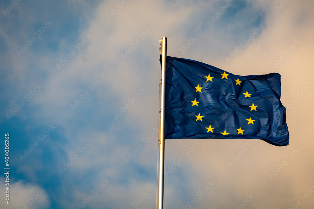 Flag of the European Union waving on a flag pole in front of a sky with clouds.