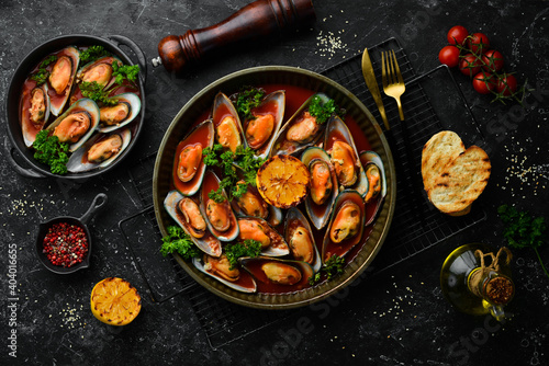 Metal tray with mussels cooked in tomato sauce with garlic, parsley and lemon. On a black stone background.