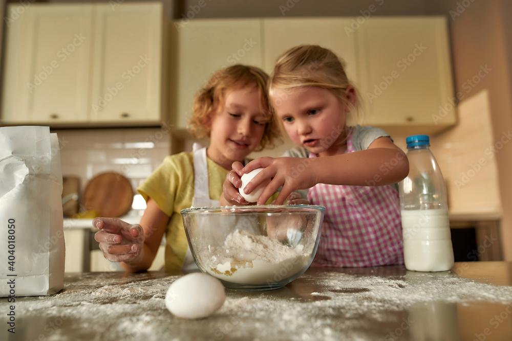 Two cute little children, boy and girl in aprons looking focused while adding eggs, preparing dough together on the kitchen table at home Two cute little children, boy and girl in aprons looking focused while adding eggs, preparing dough together on the kitchen table at home