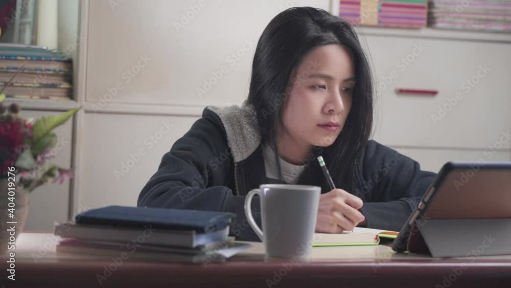 young asian female study from home by watching tablet online e-learning class lesson, hand writing with pencil on notebook, wear sweater warm night, distanced leaner, home school young asian female study from home by watching tablet online e-learning class lesson, hand writing with pencil on notebook, wear sweater warm night, distanced leaner, home school