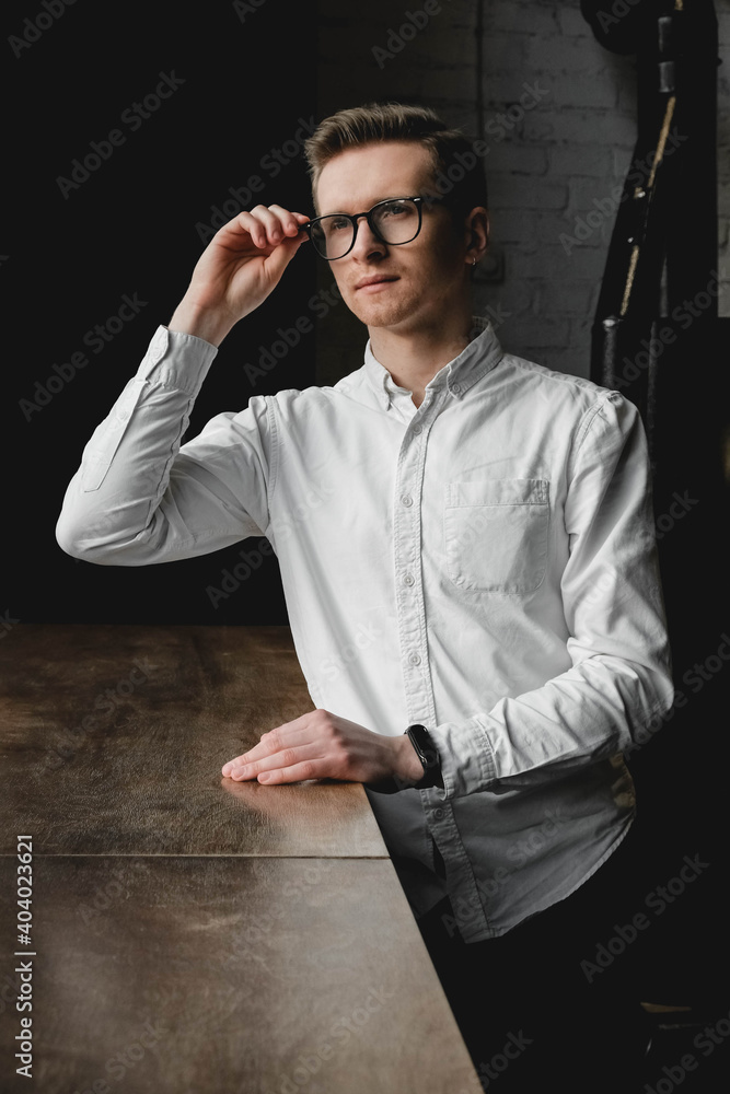 Young man in glasses and a white shirt sitting at the table on a dark background. Copy, empty space for text Young man in glasses and a white shirt sitting at the table on a dark background. Copy, empty space for text