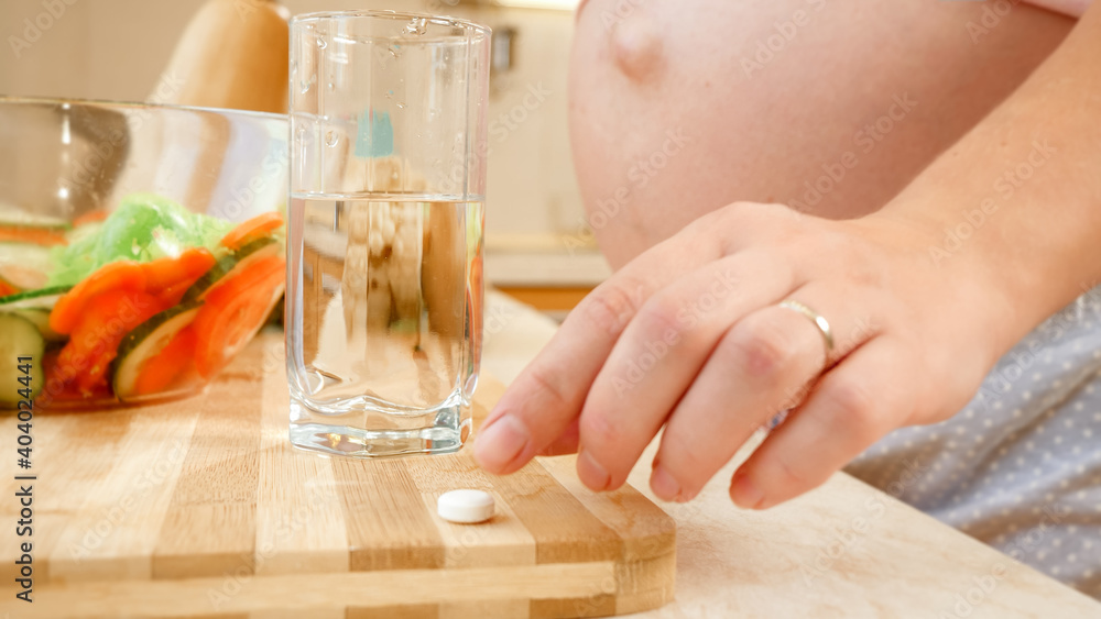 Closeup of pregnant woman taking vitamin pill and drinking glass of clear water before breakfast on kitchen. Concept of healthy lifestyle, nutrition and hydration during pregnancy Closeup of pregnant woman taking vitamin pill and drinking glass of clear water before breakfast on kitchen. Concept of healthy lifestyle, nutrition and hydration during pregnancy