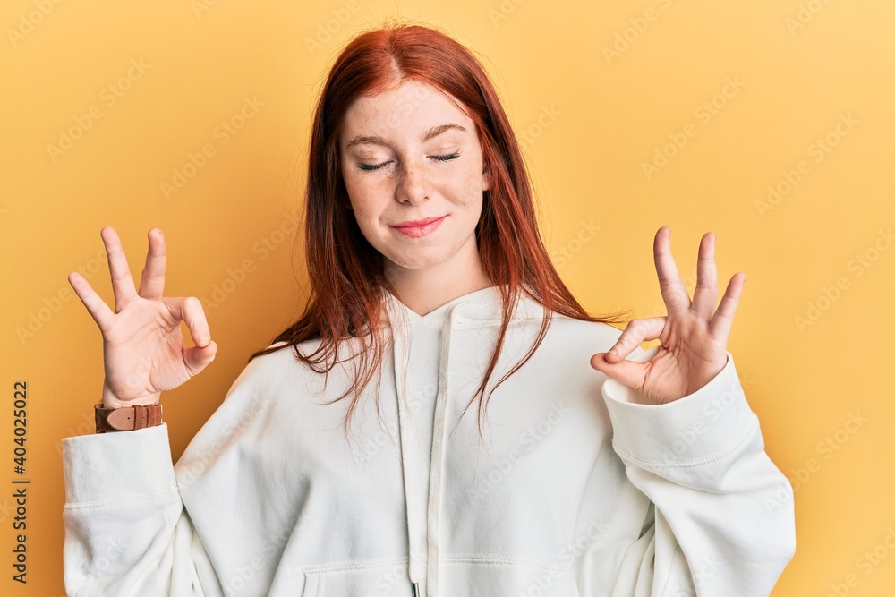 Young red head girl wearing casual sweatshirt relax and smiling with eyes closed doing meditation gesture with fingers. yoga concept. Young red head girl wearing casual sweatshirt relax and smiling with eyes closed doing meditation gesture with fingers. yoga concept.