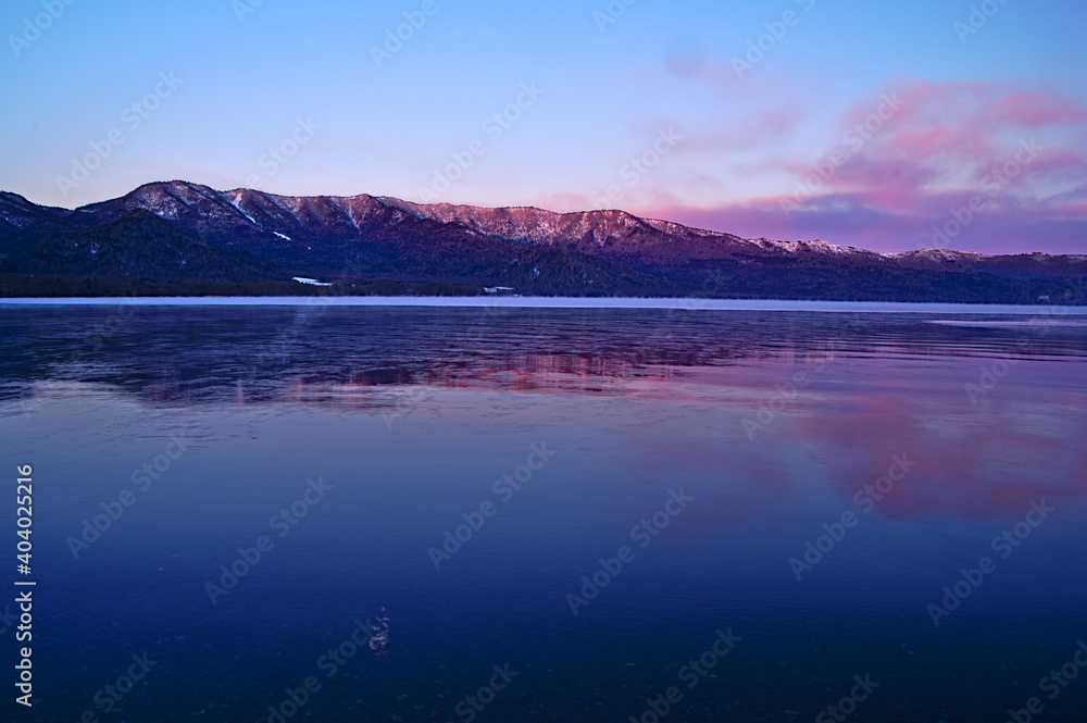 冬の湖の畔から見る夜明けの陽光に色づく山脈。