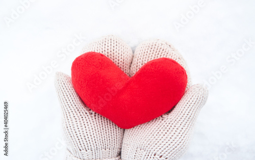 Creative greeting card for Valentines Day. Hold red soft heart toy made of fabric in beige mittens against background of white freshly fallen snow.
