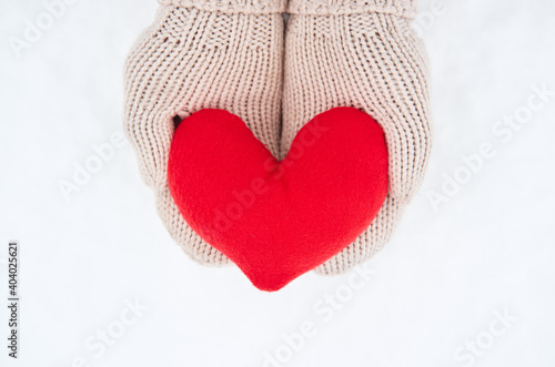 Creative greeting card for Valentines Day. Hold red soft heart toy made of fabric in beige mittens against background of white freshly fallen snow.