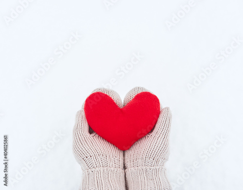 Creative greeting card for Valentines Day. Hold red soft heart toy made of fabric in beige mittens against background of white freshly fallen snow. Empty space for your greeting text.