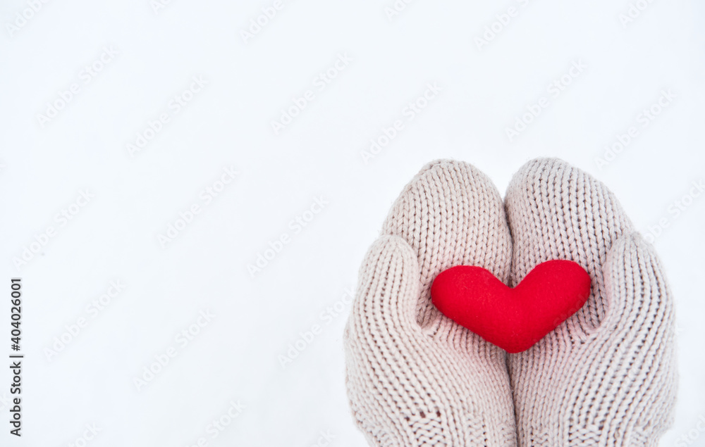 Creative greeting card for Valentines Day. Holding red little soft fabric toy heart in beige mittens against background of white freshly fallen snow. Empty space for your greeting text.