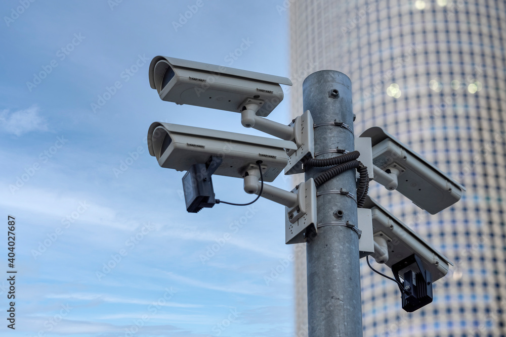 Security cameras watching from above, hinged to a street pole. Skyscraper and sky appear at the background.
