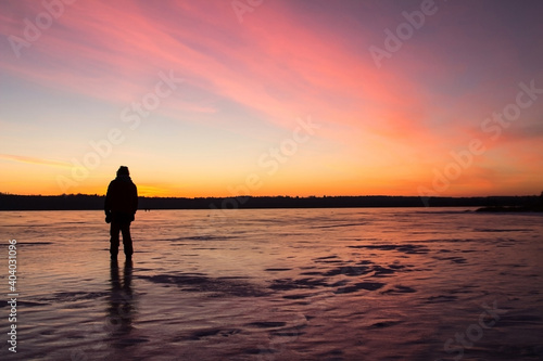 The silhouette of a man standing on the ice of a frozen lake in the light of the sunset. Winter background