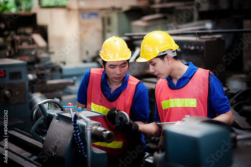 Two young asian male technicians wearing safety uniform workwear, helmets, vest and gloves are Working on a metal lathe and inspecting parts intently In industrial plants. 