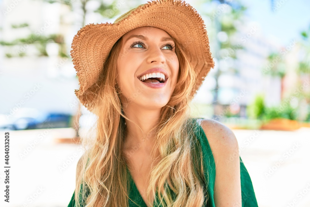 Young beautiful blonde woman on vacation wearing summer hat smiling happy. Standing with smile on face at street of city.