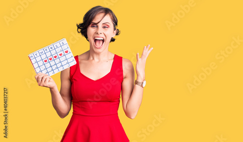 Beautiful young woman with short hair holding heart calendar celebrating victory with happy smile and winner expression with raised hands