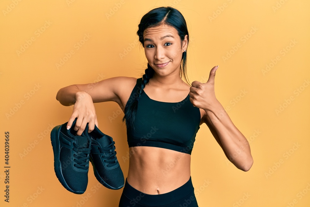 Beautiful hispanic woman wearing sportswear showing sporty shoes smiling happy and positive, thumb up doing excellent and approval sign Beautiful hispanic woman wearing sportswear showing sporty shoes smiling happy and positive, thumb up doing excellent and approval sign