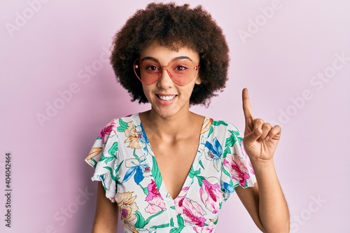 Young hispanic girl wearing summer style and hear sunglasses smiling with an idea or question pointing finger with happy face, number one