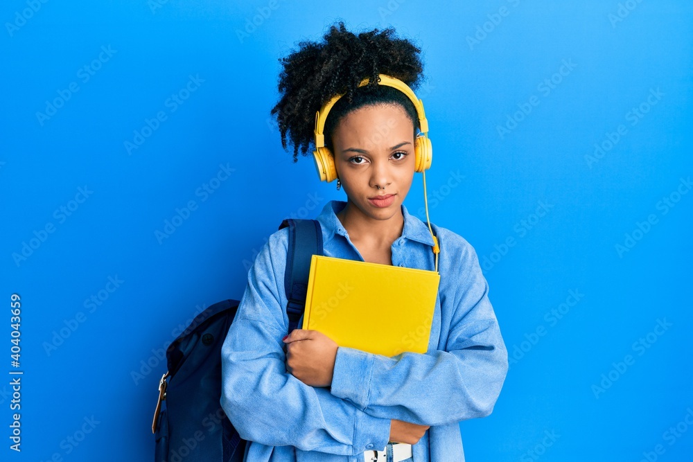 Obraz premium Young african american girl wearing student backpack and headphones holding book skeptic and nervous, frowning upset because of problem. negative person.