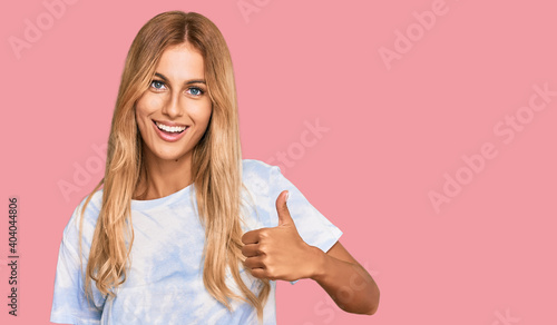 Beautiful blonde young woman wearing casual tie dye shirt smiling happy and positive, thumb up doing excellent and approval sign