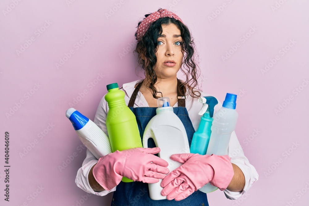 Young brunette woman with curly hair wearing cleaner apron holding cleaning products clueless and confused expression. doubt concept. Young brunette woman with curly hair wearing cleaner apron holding cleaning products clueless and confused expression. doubt concept.