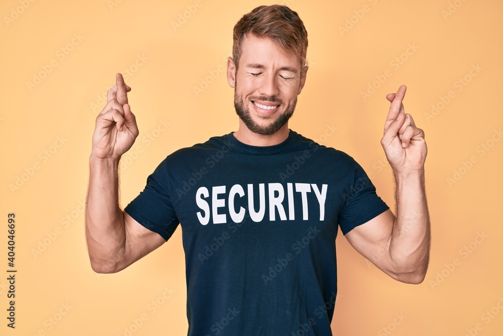 Young caucasian man wearing security t shirt gesturing finger crossed smiling with hope and eyes closed. luck and superstitious concept. Young caucasian man wearing security t shirt gesturing finger crossed smiling with hope and eyes closed. luck and superstitious concept.