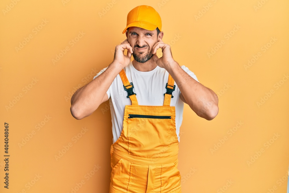 Young handsome man wearing handyman uniform over yellow background covering ears with fingers with annoyed expression for the noise of loud music. deaf concept. Young handsome man wearing handyman uniform over yellow background covering ears with fingers with annoyed expression for the noise of loud music. deaf concept.