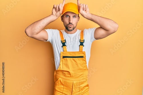 Young handsome man wearing handyman uniform over yellow background doing funny gesture with finger over head as bull horns