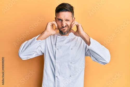 Handsome man with beard wearing professional cook uniform covering ears with fingers with annoyed expression for the noise of loud music. deaf concept. © Krakenimages.com