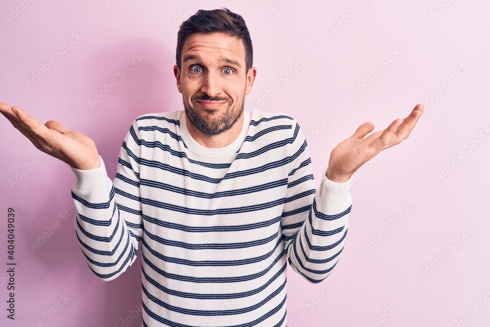 Naklejka premium Young handsome man wearing casual striped sweater standing over isolated pink background clueless and confused with open arms, no idea and doubtful face.
