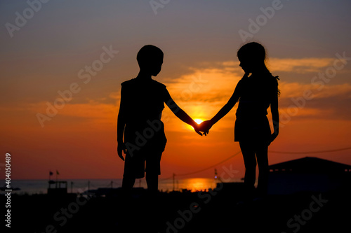 Children hold hands in the background of the sunset. Silhouettes of a young boy and girl.