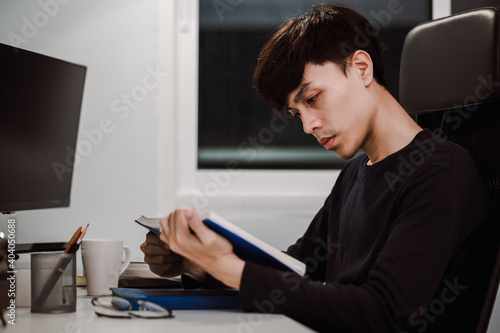 Young handsome asian man reading book at work desk late at night, Knowledge and learning concept.