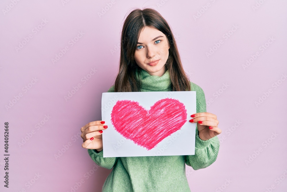 Young beautiful caucasian girl holding heart draw relaxed with serious expression on face. simple and natural looking at the camera. Young beautiful caucasian girl holding heart draw relaxed with serious expression on face. simple and natural looking at the camera.
