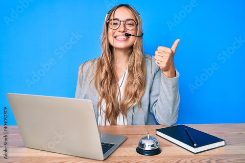 Young beautiful blonde woman wearing operator headset at the call center office smiling happy and positive, thumb up doing excellent and approval sign photo