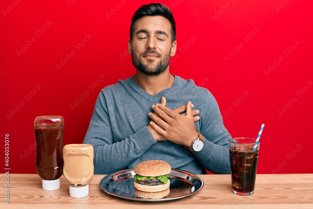 Handsome hispanic man eating a tasty classic burger and soda smiling with hands on chest, eyes closed with grateful gesture on face. health concept. Handsome hispanic man eating a tasty classic burger and soda smiling with hands on chest, eyes closed with grateful gesture on face. health concept.