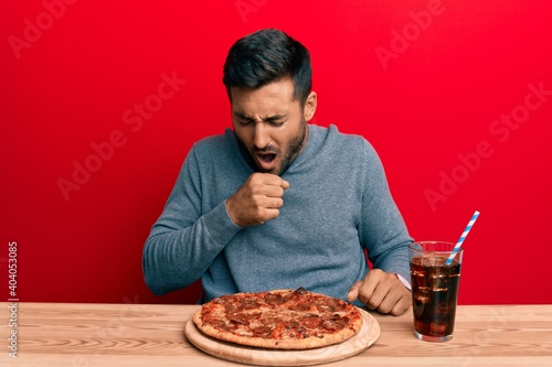 Handsome hispanic man eating tasty pepperoni pizza feeling unwell and coughing as symptom for cold or bronchitis. health care concept.
