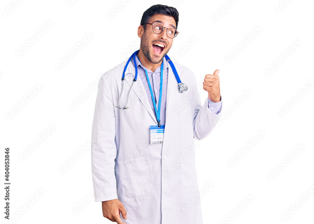 Young hispanic man wearing doctor uniform and stethoscope smiling with happy face looking and pointing to the side with thumb up. Young hispanic man wearing doctor uniform and stethoscope smiling with happy face looking and pointing to the side with thumb up.