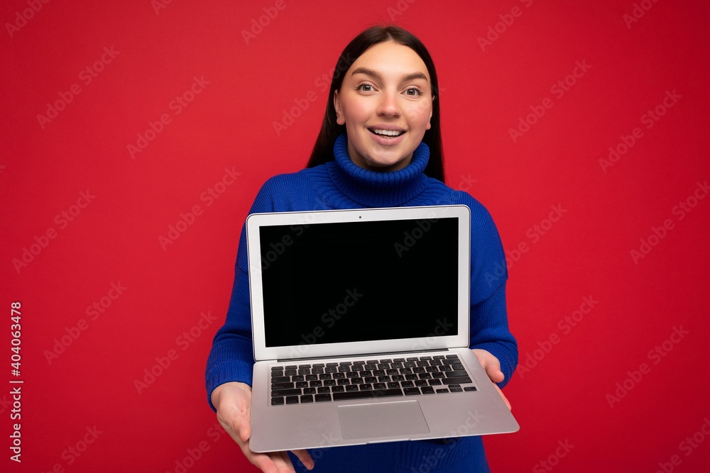 Photo of smiling charming Fascinating pretty happy beautiful young brunet woman with straight hair holding computer laptop looking at camera wearing blue sweater isolated over red wall background Photo of smiling charming Fascinating pretty happy beautiful young brunet woman with straight hair holding computer laptop looking at camera wearing blue sweater isolated over red wall background
