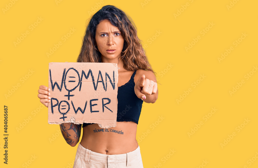 Young hispanic woman with tattoo holding woman power banner pointing with finger to the camera and to you, confident gesture looking serious Young hispanic woman with tattoo holding woman power banner pointing with finger to the camera and to you, confident gesture looking serious