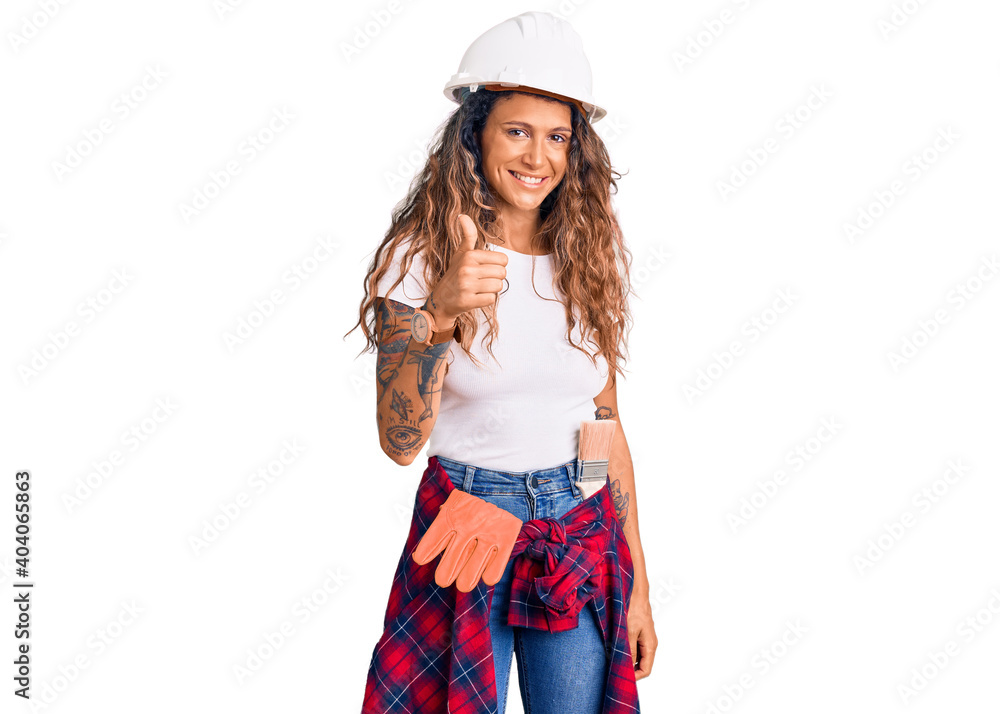 Young hispanic woman with tattoo wearing hardhat and builder clothes doing happy thumbs up gesture with hand. approving expression looking at the camera showing success. Young hispanic woman with tattoo wearing hardhat and builder clothes doing happy thumbs up gesture with hand. approving expression looking at the camera showing success.