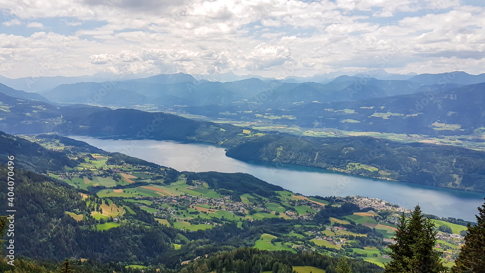 A panoramic view on the Millstaettersee lake from Granattor in Austrian Alps. The distant lake is surrounded by high mountains. Few clouds above. Endless mountain chains. Lush and vast pasture. Relax