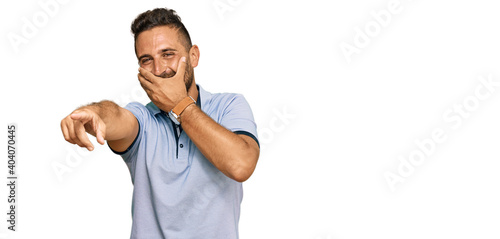 Handsome man with beard wearing casual clothes laughing at you, pointing finger to the camera with hand over mouth, shame expression