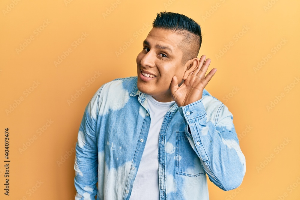 Young latin man wearing casual clothes smiling with hand over ear listening and hearing to rumor or gossip. deafness concept. Young latin man wearing casual clothes smiling with hand over ear listening and hearing to rumor or gossip. deafness concept.