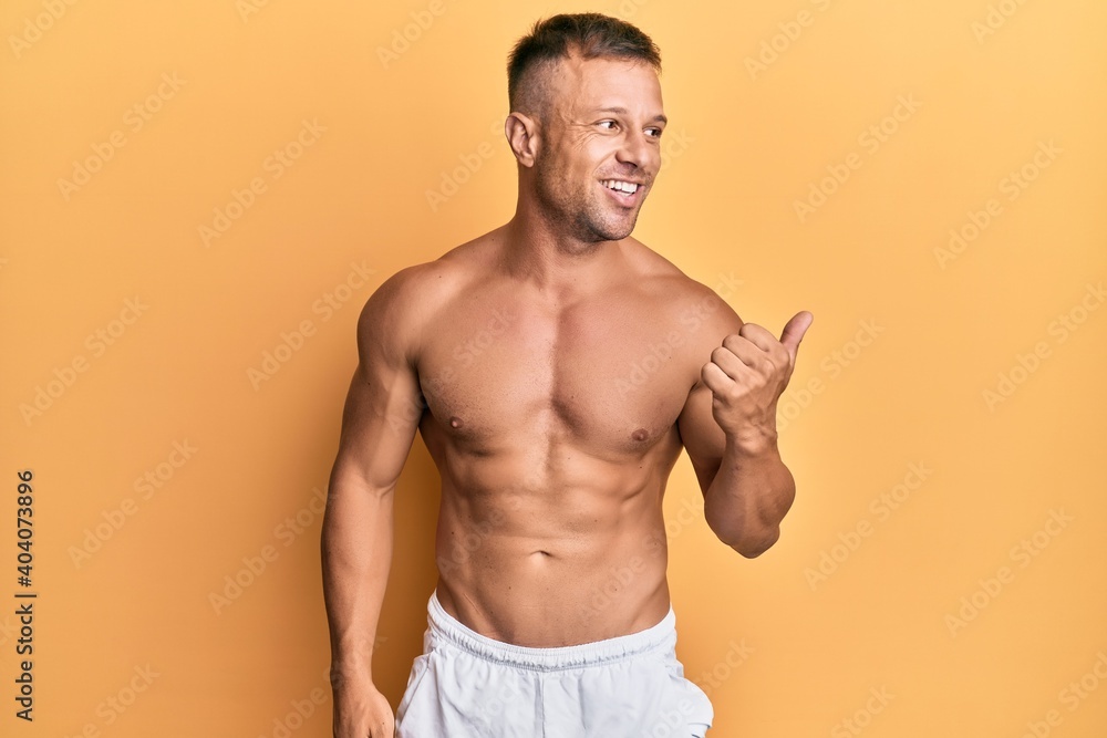 Handsome muscle man standing shirtless smiling with happy face looking and pointing to the side with thumb up.