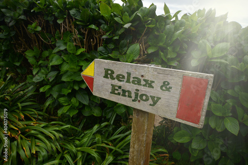vintage old wooden signboard with text relax and enjoy near the green plants.
